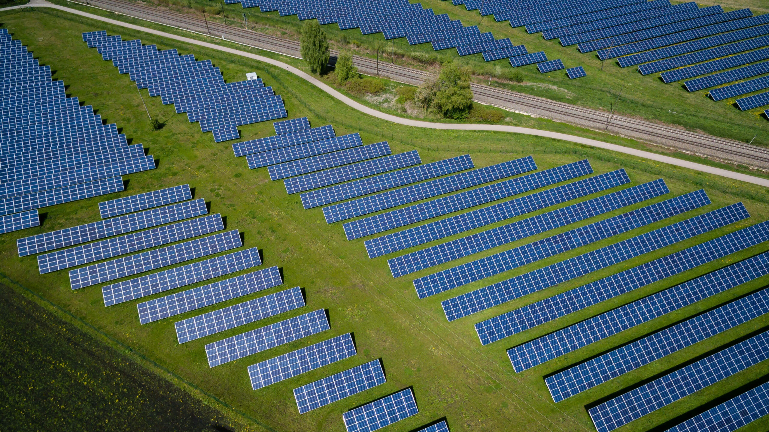 Solar farm panels in green fields, interected by a road and a railway.